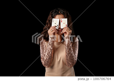 Brunette girl wearing shiny dress posing holding two playing cards in her hands standing against black studio background. Casino, poker. Close-up. 100706764