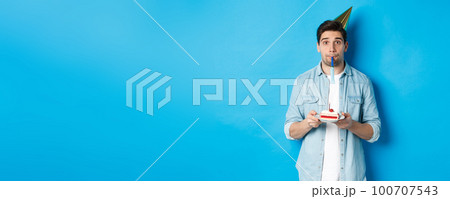 Handsome young man holding birthday cake, wearing party hat and blowing whistle, standing over blue background Handsome young man holding birthday cake, wearing party hat and blowing whistle, standing over blue background 100707543