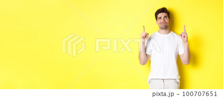 Skeptical young man pointing and looking up at something bad, judging offer, standing over yellow background 100707651
