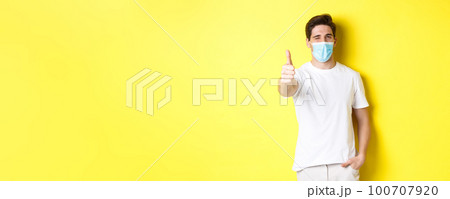 Concept of coronavirus, pandemic and social distancing. Confident young man in medical mask showing thumbs up and winking, yellow background 100707920