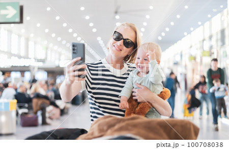 Mother taking selfie with mobile phone, while traveling with child, holding his infant baby boy at airport, waiting to board a plane. Travel with kids concept Mother taking selfie with mobile phone, while traveling with child, holding his infant baby boy at airport, waiting to board a plane. Travel with kids concept 100708038