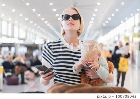Mother traveling with child, holding his infant baby boy at airport terminal, checking flight schedule, waiting to board a plane. Travel with kids concept 100708043