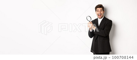 Handsome businessman in black suit, holding magnifying glass and smiling, found something, standing against white background 100708144