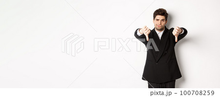 Portrait of skeptical and disappointed man in black suit, frowning upset, showing thumbs-down, dislike something bad, standing over white background Portrait of skeptical and disappointed man in black suit, frowning upset, showing thumbs-down, dislike something bad, standing over white background 100708259