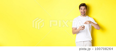 Happy man pointing at smartphone to show promo, check out internet offer, standing over yellow background Happy man pointing at smartphone to show promo, check out internet offer, standing over yellow background 100708264
