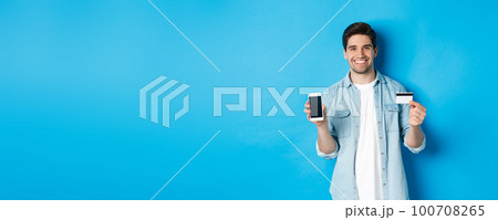 Young smiling man showing smartphone screen and credit card, concept of online shopping or banking Young smiling man showing smartphone screen and credit card, concept of online shopping or banking 100708265