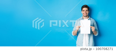 Young smiling guy in casual outfit, holding blank piece of paper with your advertisement, standing over blue background 100708271