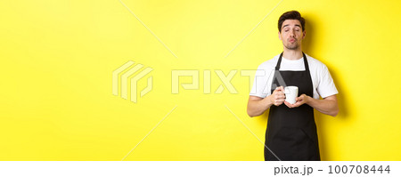 Man in black apron bringing cup of coffee and waiting for kiss, standing over yellow background 100708444
