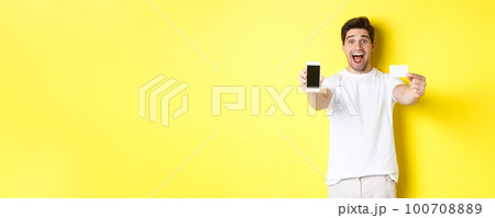 Handsome caucasian man showing smartphone screen and credit card, concept of mobile banking and online shopping, yellow background 100708889