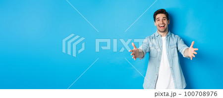 Friendly man stretching hands forward and welcome you, greeting guests or reaching for hugs, standing against blue background 100708976