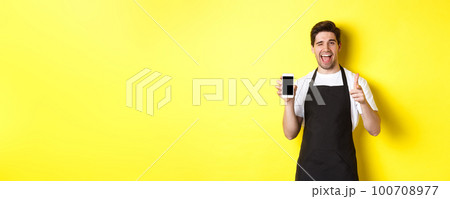 Happy waiter showing mobile screen and thumb up, recommending cafe restaurant app, standing over yellow background 100708977