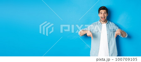 Image of excited handsome man pointing fingers down, making an announcement, standing against blue background Image of excited handsome man pointing fingers down, making an announcement, standing against blue background 100709105