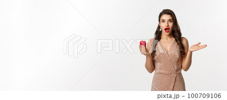 Image of amazed girlfriend holding engagement ring box, looking surprised, receive marriage proposal, wearing elegant dress, white background 100709106