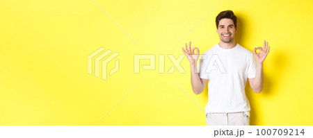 Relaxed guy smiling, showing okay signs, approve or agree, standing against yellow background 100709214