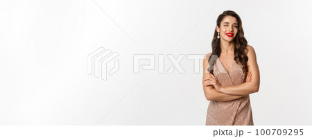 Beautiful woman in evening dress and red lipstick, winking at camera and smiling, celebrating New Year party, standing over white background Beautiful woman in evening dress and red lipstick, winking at camera and smiling, celebrating New Year party, standing over white background 100709295