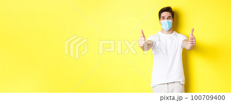 Concept of coronavirus, pandemic and social distancing. Confident man protecting himself from covid-19 with medical mask, showing thumbs up in approval, yellow background 100709400