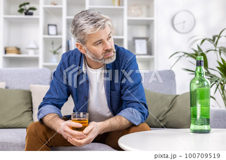 Close up of adult mature man lonely depressed drinking strong alcohol sitting on sofa in living room on day. 100709519