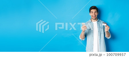 Amazed handsome man shopping online, holding mobile phone and credit card, smiling while paying for internet purchase, standing over blue background Amazed handsome man shopping online, holding mobile phone and credit card, smiling while paying for internet purchase, standing over blue background 100709937