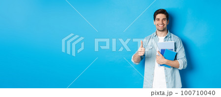 Young man student with notebooks, showing thumb up in approval, smiling satisfied, blue studio background 100710040