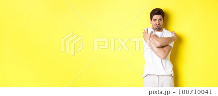 Silly and timid guy trying to comfort himself, hugging his body and frowning scared, standing over yellow background Silly and timid guy trying to comfort himself, hugging his body and frowning scared, standing over yellow background 100710041