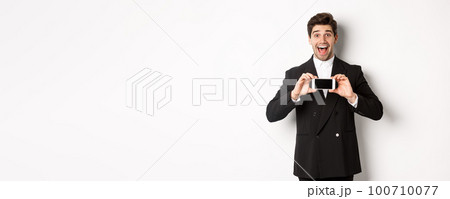 Image of cheerful, handsome man in black suit, showing smarthone screen and looking amazed, standing against white background 100710077