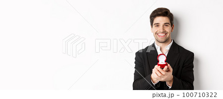 Image of handsome man looking romantic, open small box with engagement ring, making a proposal and smiling, standing against white background 100710322