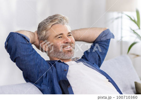 Mature adult man at home close up with hands behind head sitting on couch and thinking dreaming about future plans, gray haired person in living room at home smiling and happy with day and work. Mature adult man at home close up with hands behind head sitting on couch and thinking dreaming about future plans, gray haired person in living room at home smiling and happy with day and work. 100710360