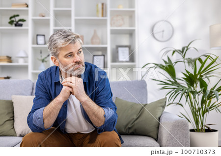 Adult mature man pensive sitting alone at home on sofa close up, gray haired person thinks looks towards window, depressed sad. Adult mature man pensive sitting alone at home on sofa close up, gray haired person thinks looks towards window, depressed sad. 100710373