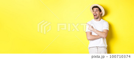 Concept of tourism and lifestyle. Excited handsome guy in straw hat checking out advertisement, pointing and looking at upper left corner logo, yellow background Concept of tourism and lifestyle. Excited handsome guy in straw hat checking out advertisement, pointing and looking at upper left corner logo, yellow background 100710374