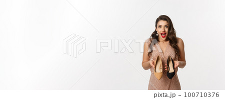 Party and celebration concept. Excited woman holding two types of heels and looking amazed, picking outfit for Christmas, standing in elegant dress over white background 100710376
