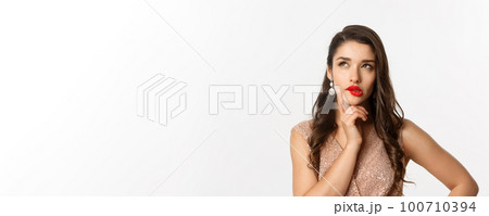 Concept of New Year celebration and winter holidays. Close-up of elegant woman with red lips and dress, looking at upper left corner and thinking, standing over white background 100710394