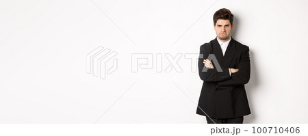 Portrait of angry handsome man in black suit, cross arms on chest and looking offended, frowning and pouting, being mad at someone, standing over white background 100710406