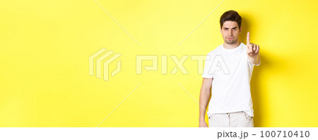 Serious man frowning displeased, shaking one finger to prohibit something, warn you, standing over yellow background 100710410