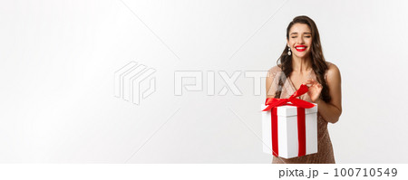 Merry Christmas. Charming woman in elegant dress open gift box and smiling delighted, unwrapping a present, standing over white background 100710549