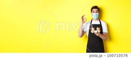 Concept of covid-19, cafe and social distancing. Barista in medical mask and black apron guarantee safety, holding takeaway cups of coffee and showing OK sign, yellow background Concept of covid-19, cafe and social distancing. Barista in medical mask and black apron guarantee safety, holding takeaway cups of coffee and showing OK sign, yellow background 100710576