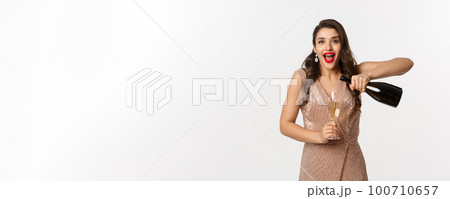 Winter holidays celebration concept. Excited woman pouring glass of champagne and staring amazed at camera, wearing party dress, standing over white background 100710657