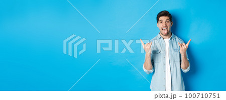 Excited handsome guy having fun, showing rock and roll sign and sticking tongue, standing against blue background 100710751