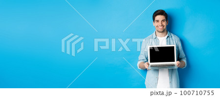 Handsome young man introduce product on laptop screen, showing computer and smiling, standing over blue background 100710755