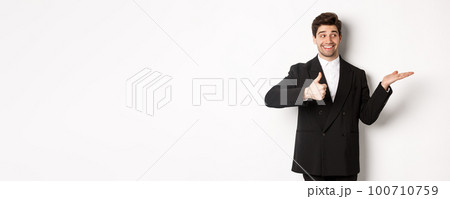 Handsome businessman in black suit, showing thumb-up and holding your product in hand over white copy space, standing against white background Handsome businessman in black suit, showing thumb-up and holding your product in hand over white copy space, standing against white background 100710759