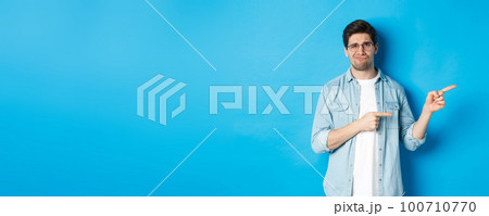 Disappointed young man in glasses pointing fingers right at copy space, showing promo banner and smirking dissatisfied, judging bad product, standing over blue background 100710770