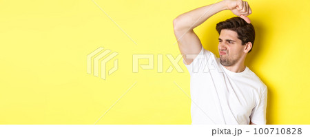 Man in sweat smelling his armpit, standing in white t-shirt and grimacing from stinky clothes Man in sweat smelling his armpit, standing in white t-shirt and grimacing from stinky clothes 100710828