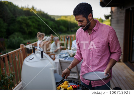 Father grilling meat and vegetable on grill during family summer garden party. 100715925