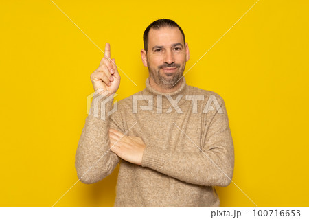 Bearded hispanic man wearing a beige turtleneck raising his index finger asking for a turn to express the great idea he had, he feels proud for having found a solution to the problem. 100716653