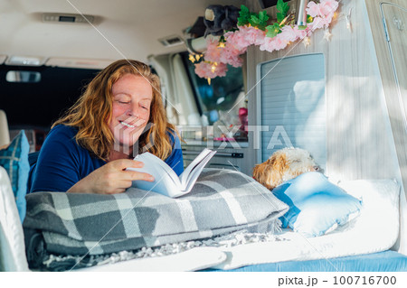 Middle-aged woman relaxing, reading book while lying in camper car vehicle with cockapoo puppy pet during road trip. Best friend. Enjoying free lifestyle, travel, vacation, freedom. Selective focus. Middle-aged woman relaxing, reading book while lying in camper car vehicle with cockapoo puppy pet during road trip. Best friend. Enjoying free lifestyle, travel, vacation, freedom. Selective focus. 100716700