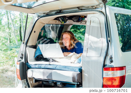Smiling middle-aged woman working on laptop while lying in camper car vehicle with cockapoo puppy pet during road trip. Remote worker people. Enjoying free lifestyle, travel, vacation, freedom. Smiling middle-aged woman working on laptop while lying in camper car vehicle with cockapoo puppy pet during road trip. Remote worker people. Enjoying free lifestyle, travel, vacation, freedom. 100716701