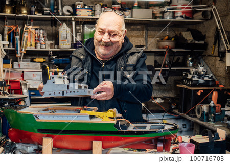 Senior man, pensioner making ship model building handcraft on table with different materials in his garage. Retired elderly man boat modeling hobby. Active Retirement lifestyle. Selective focus. Senior man, pensioner making ship model building handcraft on table with different materials in his garage. Retired elderly man boat modeling hobby. Active Retirement lifestyle. Selective focus. 100716703