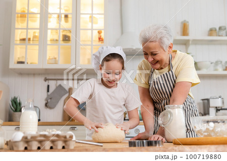 Happy family in kitchen. Grandmother and granddaughter child cook in kitchen together. Grandma teaching kid girl knead dough bake cookies. Household teamwork helping family generations concept 100719880