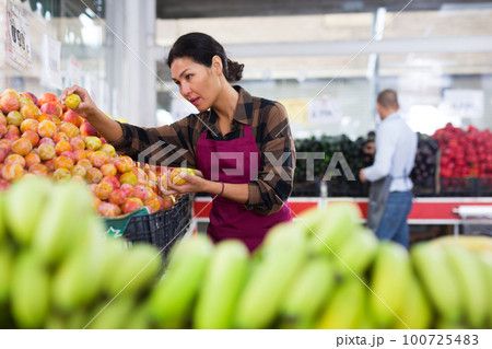 Woman in apron stacking plums in greengrocer 100725483