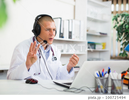 Doctor in earphones having online consultation with patient on laptop Doctor in earphones having online consultation with patient on laptop 100725494