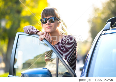 Young woman driver enjoying warm summer day standing beside her car on city street. Travelling and vacation concept. Young woman driver enjoying warm summer day standing beside her car on city street. Travelling and vacation concept. 100735250